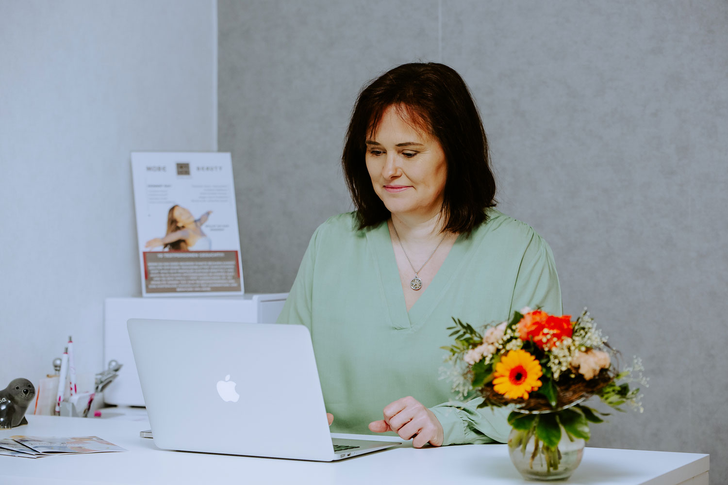 Frau mit brauner Haarfarbe, lächelt, am Laptop sitzend, grünem Oberteil, Blumenarrangement am Tisch.