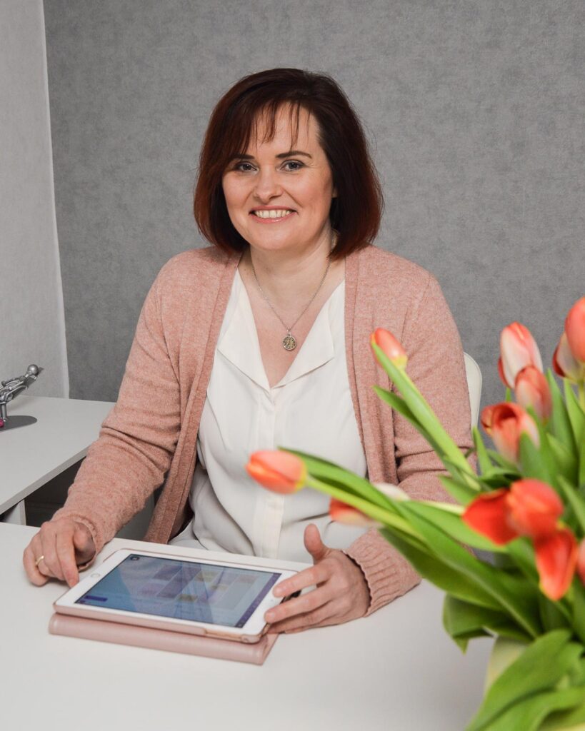 Eine lächelnde Frau sitzt an einem Tisch mit einem Tablet und Tulpen.